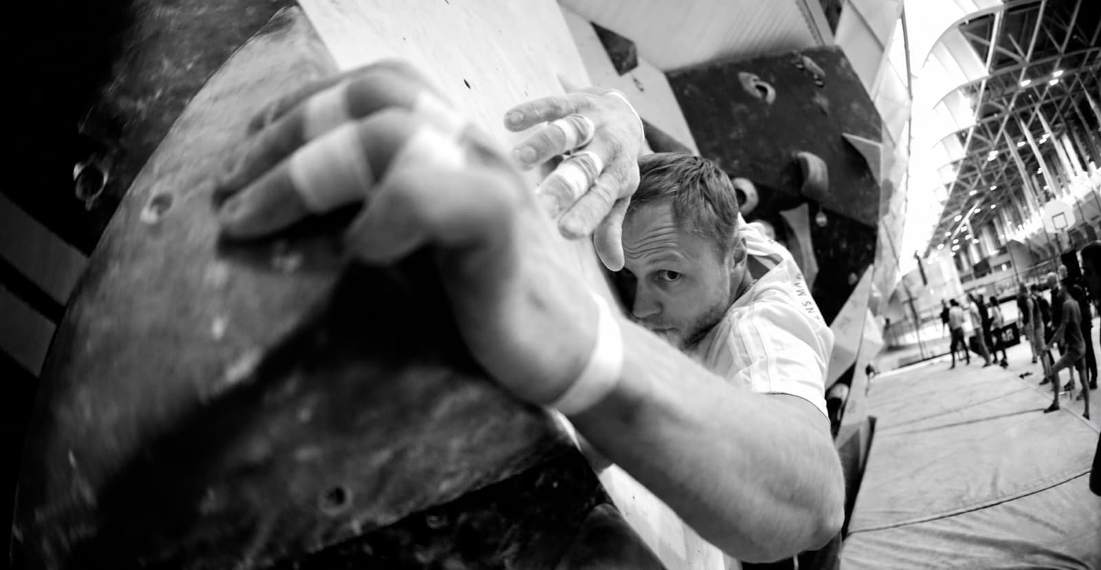 Climber with taped fingers gripping a large volume on an indoor bouldering wall, black and white fisheye shot.